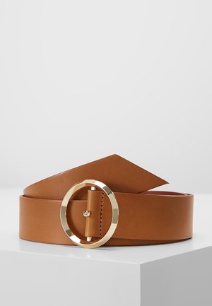 Ceinture en cuir marron clair avec une boucle circulaire dorée brillante, enroulée et reposant sur une surface blanche sur un fond uni.