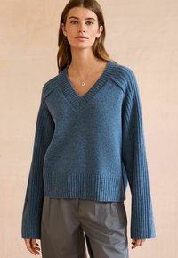 Blå, strikket pullover sweater med V-hals, ribbede manchetter og kant. Stoffet ser blødt ud med en tekstureret finish. Bæres med grå bukser.