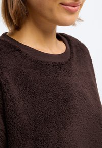 Brūns vilnas krekls ar mīkstu, teksturētu virsmu un apaļu kakla izgriezumu. Audums izskatās silts un pūkains.