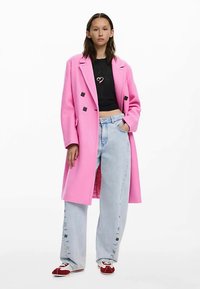 Rosa yllekappa med dubbelknäppt design, svarta knappar, kombinerad med ljusblå vida jeans och röda och vita sneakers.