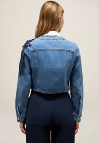 Chaqueta de denim recortada en azul claro con detalles decorativos de cuentas en un hombro, que cuenta con mangas largas y puños abotonados.