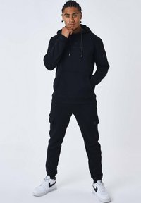 Felpa nera con cappuccio e pantaloni cargo in tessuto morbido, dotati di tasca frontale, cordini e tasche laterali. Indossati con sneakers bianche.