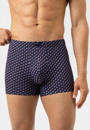 Uomo che indossa boxer blu navy con motivo di bicchieri da cocktail rosa, che aggiusta la cintola con la mano sinistra.