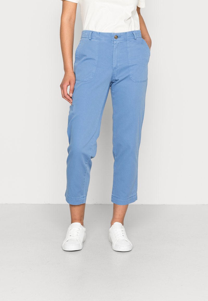 Femme portant un pantalon court bleu clair avec des poches et des baskets blanches, debout devant un fond clair.