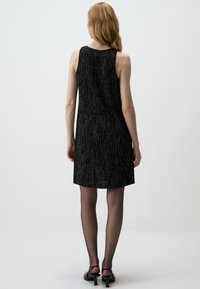 Schwarzes ärmelloses Kleid mit strukturierter Oberfläche, das subtile metallische Fäden für zusätzliche Dimension aufweist, oberhalb des Knies endet und mit transparenten Strumpfhosen kombiniert wird.