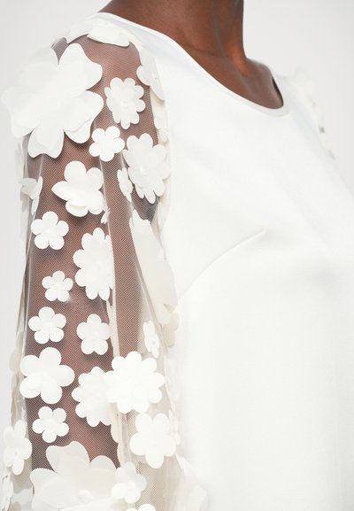 Blusa blanca con mangas de tul bordadas con flores, que presenta aplicaciones florales texturizadas sobre malla. Cuerpo liso y escote redondo.