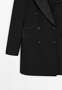 Schwarzer zweireihiger Blazer mit einem Satin-Revers, ausgestattet mit vier Knöpfen und einer Seitentasche. Glatte Textur mit einem maßgeschneiderten Design.