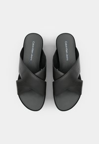 Černé pantofle na klínku vyrobené z hladkého syntetického materiálu s propleteným designem a plochými podrážkami. Na stélce je logo "Calvin Klein Jeans".