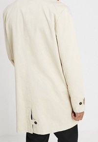 Beige pitkähihaisella takilla, jossa on nappisulkijat ja selkäventtiili, käytössä olevan henkilön takaa otettu kuva.