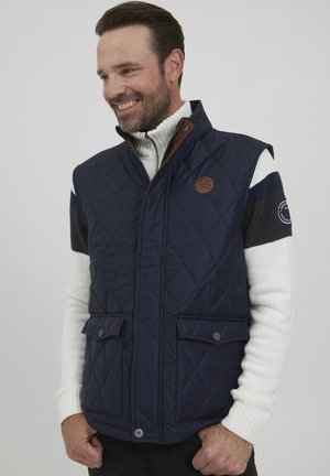 Marineblauwe gewatteerde gilet met een hoge kraag, bruine leren accenten en zijzakken. Gedragen over een witte geribde trui met marineblauwe en grijze strepen.