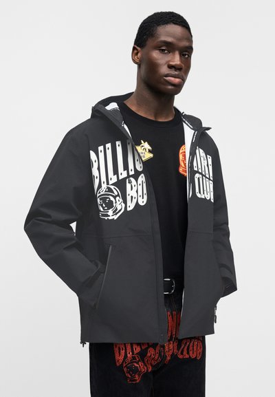 Μαύρο αδιάβροχο μπουφάν με κουκούλα, με μεγάλα λευκά γράμματα και γραφικό αστροναύτη. Φορεμένο πάνω από μαύρο πουκάμισο με πολύχρωμα μπαλώματα.