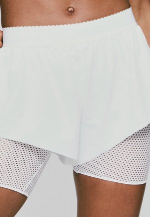 Witte sportshorts met een geschulpte tailleband over witte mesh leggings met een klein, gelijkmatig verspreid gaatjespatroon.