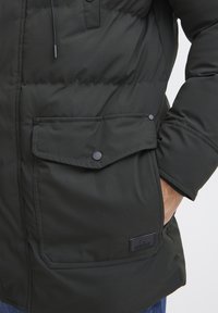 Chaqueta de plumas verde oscuro con cremallera frontal, botones a presión en el bolsillo del pecho y una etiqueta negra texturizada en el lado inferior.