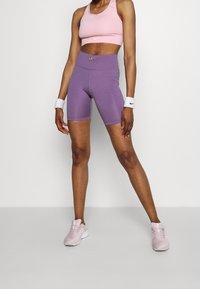 Vaaleanpunainen urheiluliivi ja korkeavyötäröiset violettit shortsit, molemmat istuvat hyvin, yhdistettynä vaaleanpunaisiin urheilukenkiin. Valkoiset rannekkeet tuovat kontrastia.