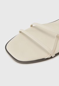 Minimalist flat sandal dengan bagian atas berwarna krim lembut, menampilkan dua tali tipis dan outsole hitam yang sedikit membulat.