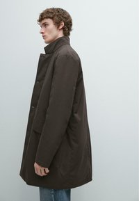 Brauner Mantel mit hohem Kragen, Druckknöpfen, Vorderentaschen und lockerem Schnitt; hergestellt aus glattem, strapazierfähigem Stoff.