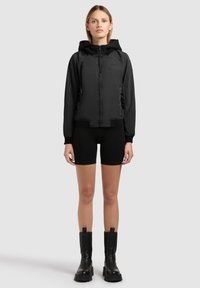 Ung kvinde står iført en sort hættejakke med lynlås, sorte cykelshorts og sorte kraftige støvler til midt på læggen på en ensartet lys baggrund.