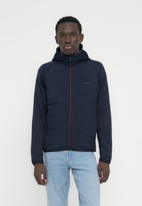 Νέος άνδρας που φοράει μπλε σκούρο μπλε μπουφάν με καπό και φερμουάρ μπροστά και ανοιχτόχρωμο τζιν, στέκεται μπροστά σε λευκό απλό φόντο.