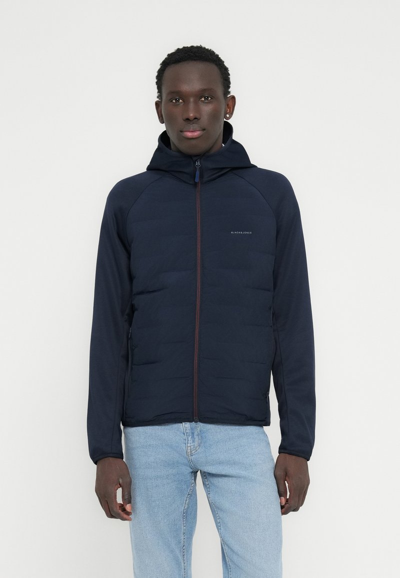 Νέος άνδρας που φοράει μπλε σκούρο μπλε μπουφάν με καπό και φερμουάρ μπροστά και ανοιχτόχρωμο τζιν, στέκεται μπροστά σε λευκό απλό φόντο.