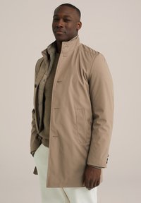 Abrigo impermeable beige con un alto cuello, botones frontales y bolsillos laterales, superpuesto a un suéter de punto marrón y pantalones blancos.