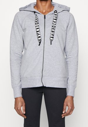 Personne portant un sweat à capuche zippé gris clair avec des cordons de serrage à motifs noir et blanc et un pantalon noir sur un fond uni.