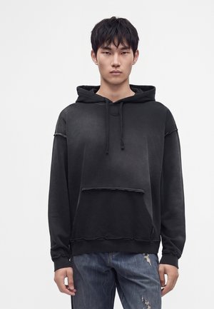 Giovane uomo con capelli corti e neri che indossa una felpa nera con cappuccio e jeans blu strappati, in piedi davanti a uno sfondo bianco uniforme.