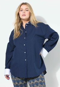 Granatowa koszula na guziki z długim rękawem w pionowe białe paski, z kontrastującymi białymi mankietami i luźnym krojem; zestawiona z wzorzystymi spodniami.