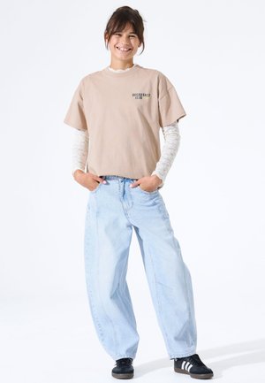 T-shirt en coton brun clair avec le texte « BREAKFAST CLUB » ; superposé avec un haut à manches longues en dentelle blanche. Jean large bleu clair et baskets noires.