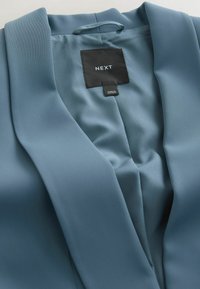 Niebieska, dopasowana marynarka z czarną metką marki "NEXT" oraz metką "Made in Vietnam" na gładkiej podszewce.