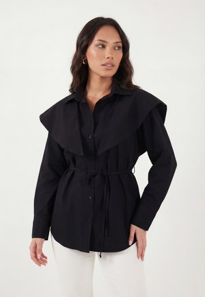 Kvinde med langt mørkt hår iført en sort knapbluse med vid krave og taljebånd, kombineret med hvide bukser mod en ensfarvet baggrund.