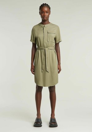 Robe-shirt vert olive avec une poche poitrine, des manches courtes, une taille ceinturée et un ourlet arrondi. Associée à des chaussures noires à enfiler.