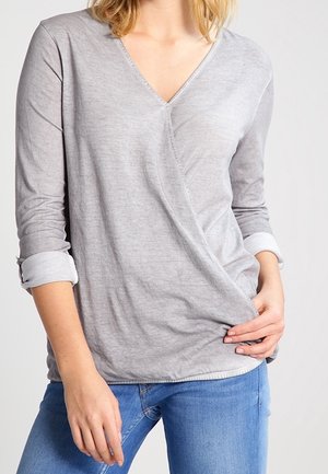 Femme portant un haut gris clair à manches longues avec col en V et poignets retournés, et un jean bleu, tenant l’ourlet du haut d’une main.