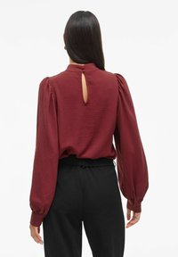 Blusa bordeaux con maniche lunghe a sbuffo, tessuto texturizzato e chiusura con bottone sul retro. Indossata con pantaloni neri su misura. Design semplice ed elegante.