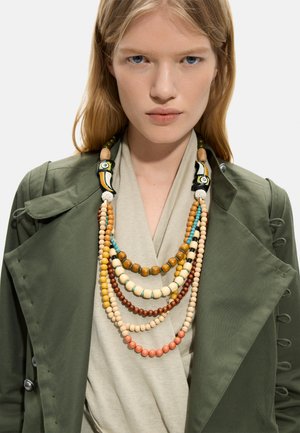 Kobieta z długimi blond włosami nosi warstwowy, wielokolorowy naszyjnik z koralików z dwoma czarno-żółtymi koralikami w kształcie tukana, oliwkowo zieloną kurtkę i beżowy top.