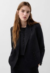 Jeune femme aux longs cheveux châtain clair portant un blazer texturé noir et un haut assorti, se tenant devant un fond clair.