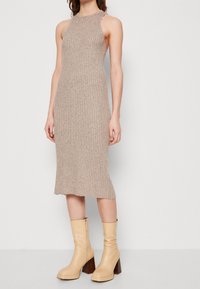 Stickad, ärmlös, beige klänning med ribbad textur och en fåll som böjer sig lätt. I kombination med beige h öga läderstövlar.