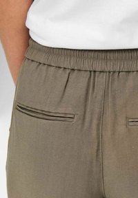 Gros plan sur un pantalon kaki à taille élastique avec poches passepoilées arrière, porté avec une chemise blanche.