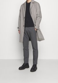 Grå rutigt trenchcoat över svart strukturerad skjorta, kombinerat med smala grå byxor och svarta snörkängor. Sammansatt tyg, skräddarsydd passform.