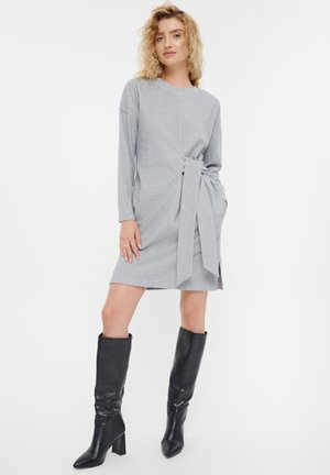 Grijze jurk met lange mouwen, een geknoopte taille-detail en zijsplitten. Gecombineerd met knielange zwarte leren laarzen. Soepele stoftextuur.