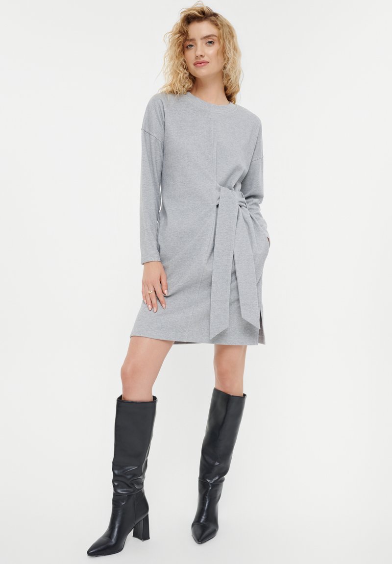 Vestido gris con mangas largas, detalle de cintura anudada y aberturas laterales. Combinado con botas de cuero negras hasta la rodilla. Textura de tela suave.