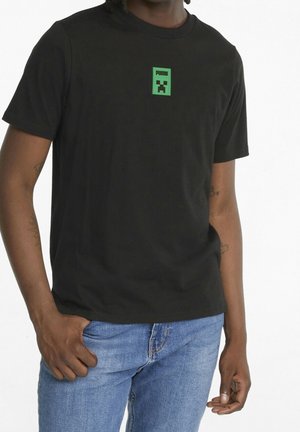 Černé bavlněné tričko s grafickým motivem zelené pixelované postavy na hrudi a kulatým výstřihem. Nosené s modrými džíny.