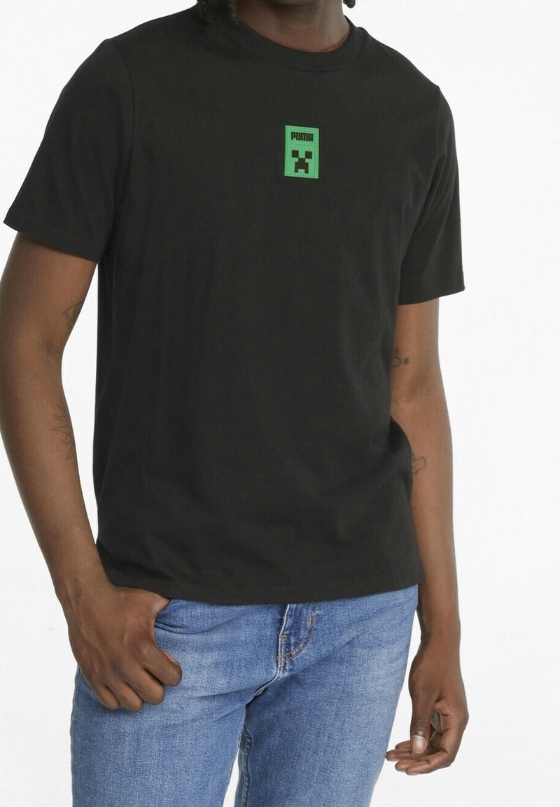 T-shirt en coton noir avec un graphisme de personnage pixelisé vert sur la poitrine et un col rond. Porté avec un jean bleu.