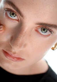 Primer plano de un rostro con piel clara, ojos verdes y cejas definidas. Color de labios natural, lleva un pendiente de aro dorado. Fondo desenfocado en blanco.