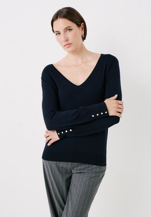 Maglione navy a V con polsini a costine e dettagli in bottone dorato, realizzato in tessuto morbido, abbinato a pantaloni grigi a righe.