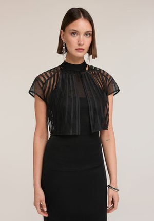 Donna con capelli corti castani che indossa un crop top nero trasparente a righe sopra un vestito nero, orecchini e braccialetti d'argento, rivolta in avanti.