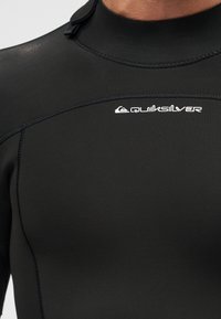 Κοντινό πλάνο ενός ατόμου που φοράει μαύρη στολή καταδύσεων Quiksilver, δείχνοντας το πάνω μέρος του στήθους και την περιοχή του λαιμού με ορατές ραφές και λογότυπο.