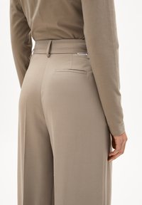 Beige maßgeschneiderte Hose mit glatter Textur, ausgestattet mit einer Gesäßtasche und einem taillierten Bund. Der Stoff wirkt leicht und strukturiert.