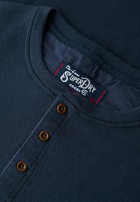 Navyblaue Henley-Bluse aus weichem Stoff, mit rundem Ausschnitt, vier braunen Knöpfen und einem innen angenähten Superdry-Label.