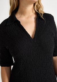Polo shirt noir texturé avec un col en V et des manches courtes. Le tissu a un aspect froissé, créant un détail de surface unique.