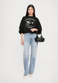 Mulher a usar um suéter preto de gola redonda com um gráfico de Cupido branco e texto, jeans azul claro com manchas de tinta rosa, segurando uma mala preta e sapatos de salto brancos.